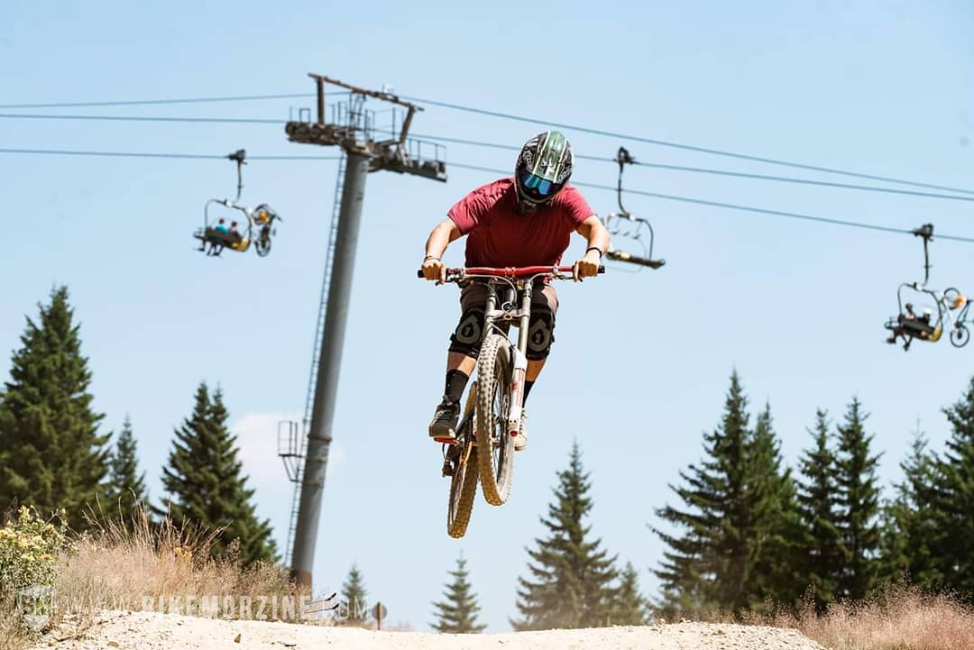 Chris airborne on a mountain bike at a bike park, a chairlift and pine trees behind.