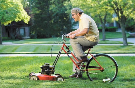 A man pedalling a bicycle bolted to a lawnmower, cutting a stripe across a suburban lawn.