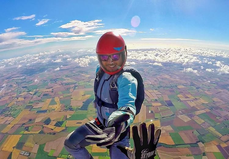 Chris skydiving in a red helmet, reaching toward the camera high above patchwork fields.