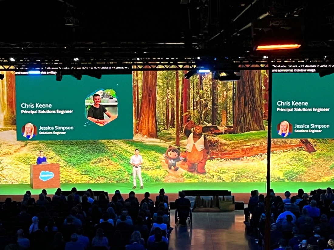 Chris on a large conference stage next to a co-presenter, his name and 'Principal Solutions Engineer' shown on the big screens.